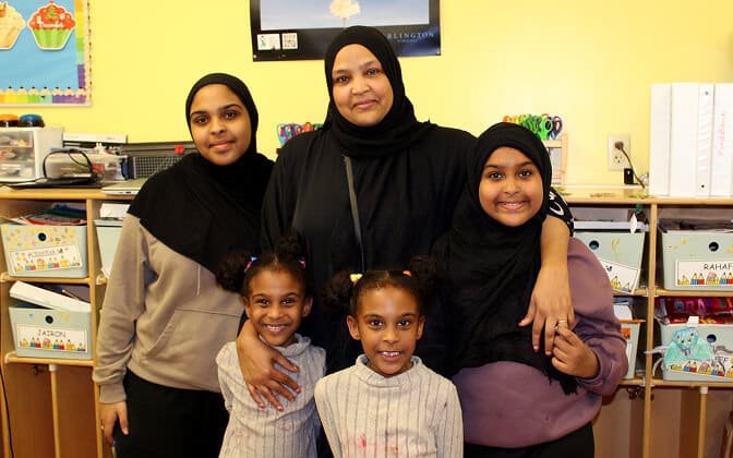 A woman wearing a black hijab stands smiling with her arms around four girls in a classroom, embodying Investing in the Future: One Family’s Path Through AHC’s Programs, with shelves, binders, and children’s artwork in the background.