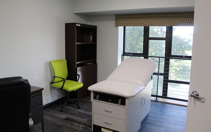 A small medical exam room with a white exam table, a bright green chair, a desk, a bookshelf, and a large window letting in natural light.