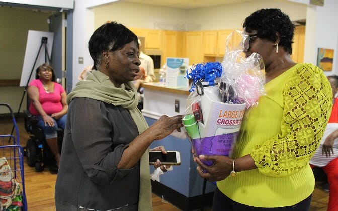 Two women stand indoors, one holding a decorated gift basket. The woman on the left gestures towards the basket and holds a phone. Other people are seated in the background in a casual setting.