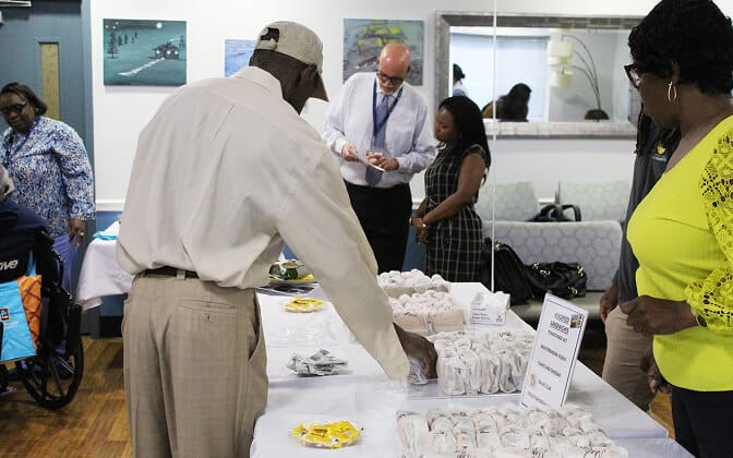 Several people stand around a table with neatly arranged snacks and informational materials in a brightly lit room. One man in a cap reaches for an item, while others chat and look on. Paintings and a mirror hang on the walls.