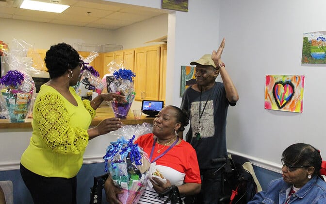 A woman in a yellow top hands a gift basket to a smiling seated woman, while a man raises his hand and another woman sits nearby. Colorful artwork decorates the walls in the background.