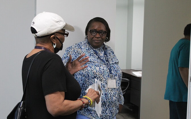 Two women stand indoors having a conversation; one wears a white cap, black mask, and black shirt, while the other, in glasses and a blue patterned blouse, listens attentively. A third person stands partially visible in the background.