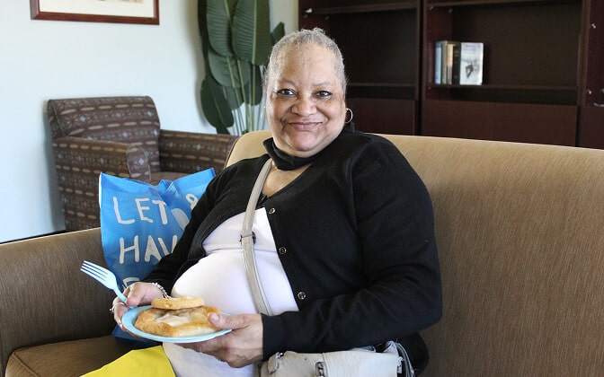 A smiling woman sits on a couch holding a plate of pastries. She is wearing a black cardigan, white shirt, and yellow skirt. A blue bag with the words LETS HAVE is in the background.