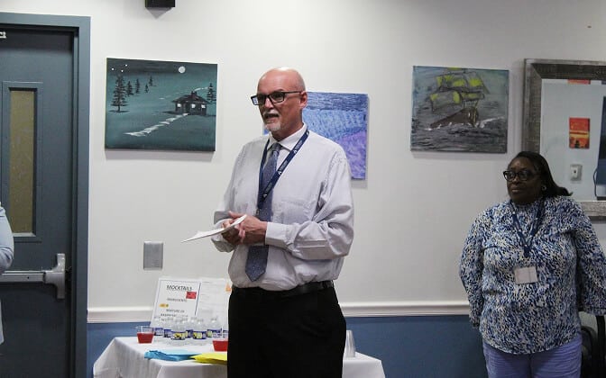 A man in a shirt and tie speaks while holding papers, standing next to a woman with glasses. They are indoors with paintings on the wall behind them and a table with snacks and drinks nearby.