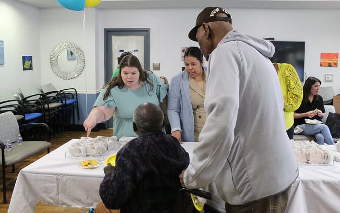 A woman behind a table with wrapped food items points while speaking to a man in a cap and another man in a wheelchair. Another woman stands beside her; others are seated in the background. The scene appears to be at a community event.