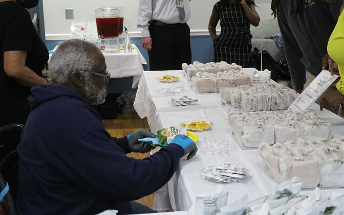 A man wearing gloves and a mask sits at a table filled with wrapped sandwiches and snacks, serving food to people at an indoor event. Other people stand around the tables, and a drink dispenser is visible in the background.