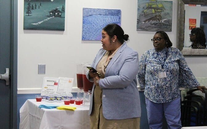 Two women stand near a table with drinks and a sign labeled “Mocktails.” The table is set against a wall decorated with paintings, and one woman is holding a phone while looking to her left.