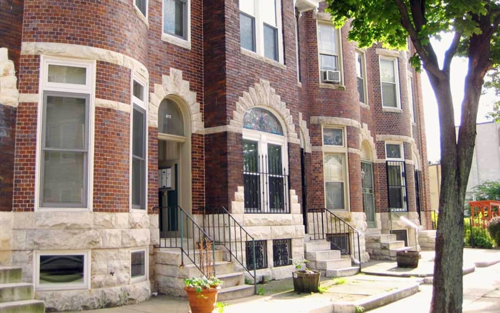 A row of brownstone townhouses with arched doorways, stone foundations, black railings, and steps leading to each entrance. A tree and potted plants are visible in the foreground on a sunny day.