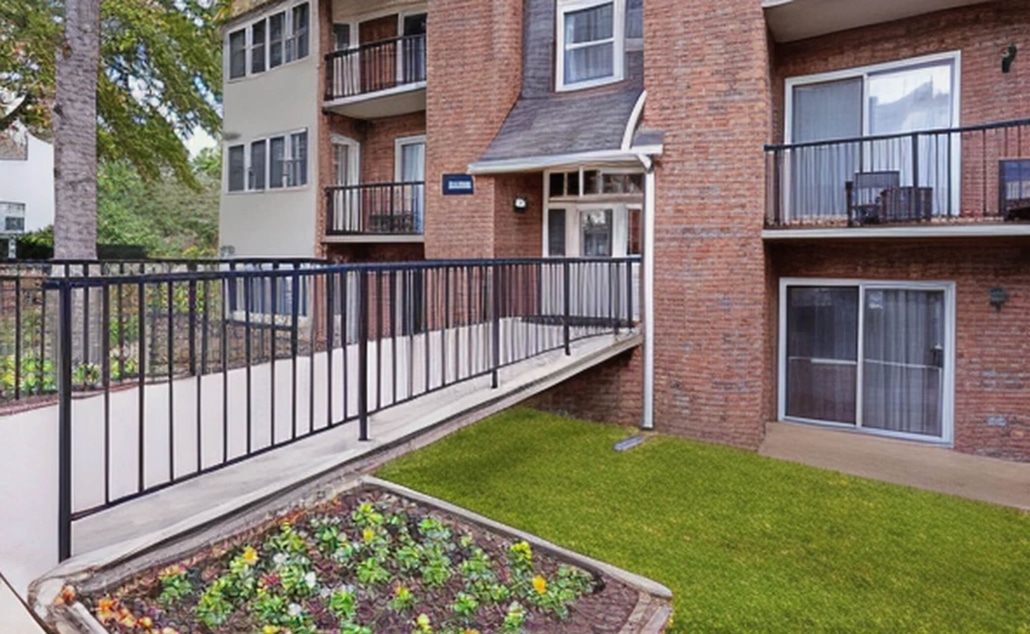 A brick apartment building with balconies, large windows, a ramp with black railings for accessibility, a garden bed with flowers, and a small grassy area in front.