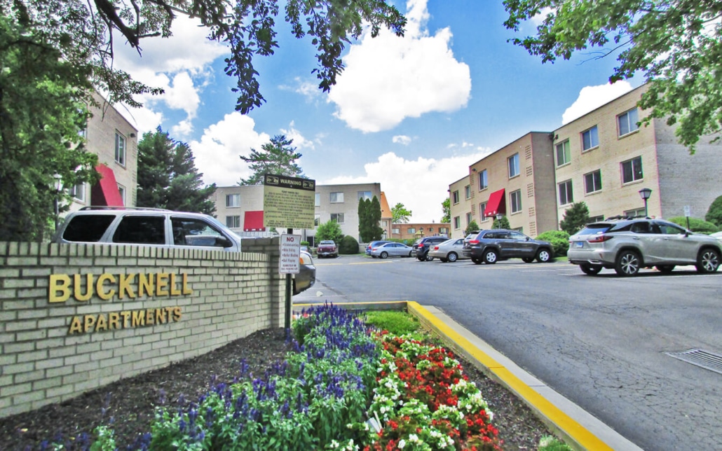 A brick sign reading Bucknell Apartments stands by a driveway leading to several tan apartment buildings. Colorful flowers are in the foreground, and parked cars line the lot under a partly cloudy sky.