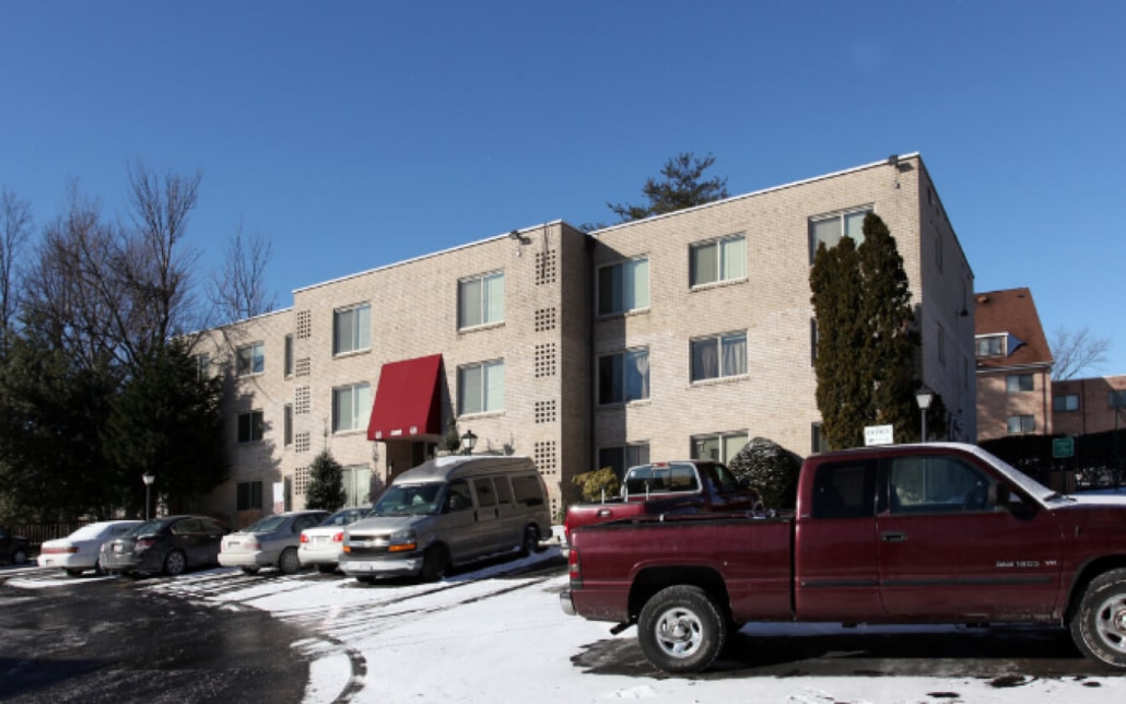 A three-story apartment building with a red awning over the entrance, several parked cars, and snow covering the ground under a clear blue sky.