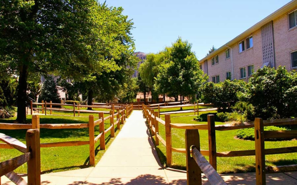 A sunlit sidewalk runs between grassy lawns and trees, bordered by wooden fences and flanked by apartment buildings on the right. The sky is clear and blue, creating a peaceful, residential atmosphere.