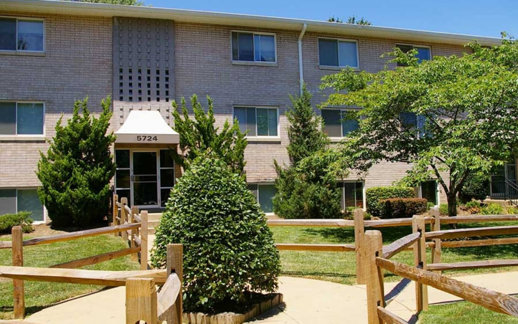 A three-story brick apartment building with large windows and a central entrance marked 5724. There is a landscaped walkway, wooden fencing, bushes, and trees in front of the building on a sunny day.