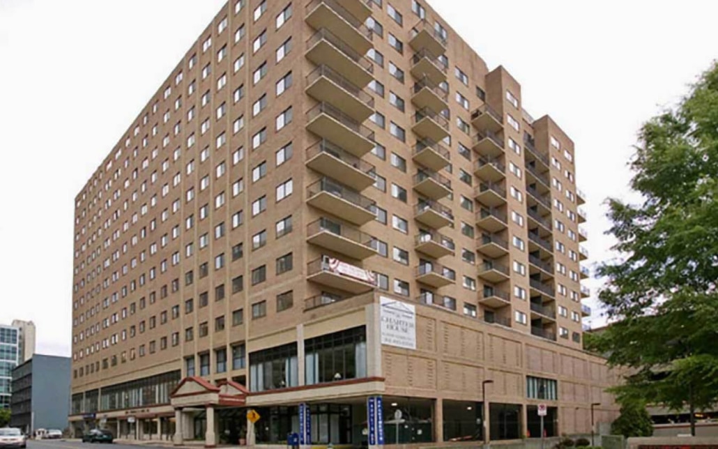 A large multi-story apartment building with a brick exterior, rows of balconies, and numerous windows, located on a city street corner with some trees and other buildings nearby.