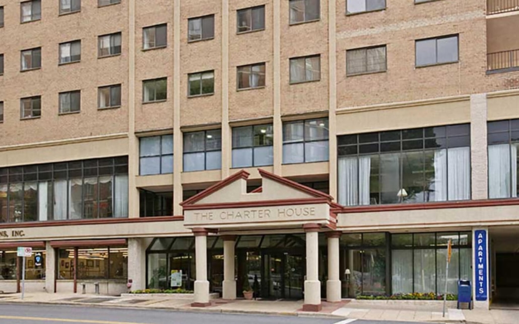 The image shows the entrance of The Charter House, a multi-story brick apartment building with large windows and a covered entryway. A blue sign reading Apartments stands near the sidewalk.