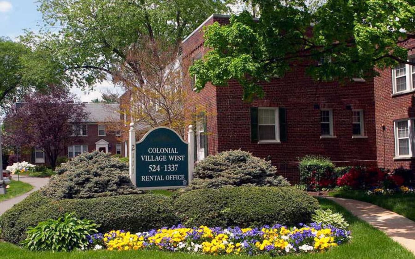 A well-kept brick apartment complex with green lawns, blooming flowers, and a sign reading Colonial Village West Rental Office 524-1337. Trees and shrubs line a winding sidewalk in a residential area.