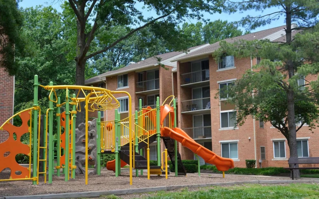 A colorful playground with slides, climbing structures, and monkey bars stands on mulch in front of a brick apartment building surrounded by green trees.