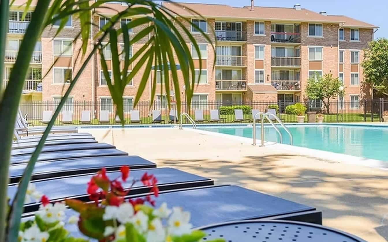 A swimming pool with lounge chairs and a table with flowers in the foreground, surrounded by a fence and an apartment building with balconies in the background. Greenery and trees are also visible.