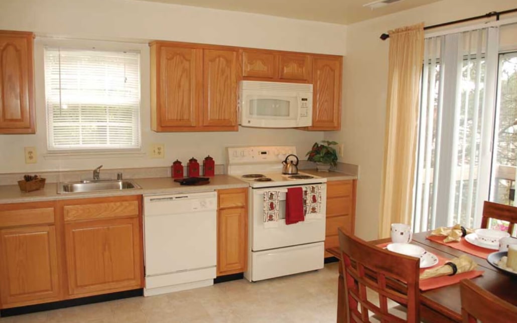 A bright kitchen with wooden cabinets, white appliances including a stove, microwave, and dishwasher, a sink under a window, and a small dining table set for two near sliding glass doors with light curtains.