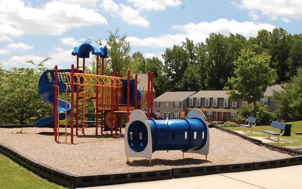 A colorful playground with slides, climbing structures, and a blue tunnel on a gravel surface is surrounded by grass and trees, with benches and houses in the background under a partly cloudy sky.