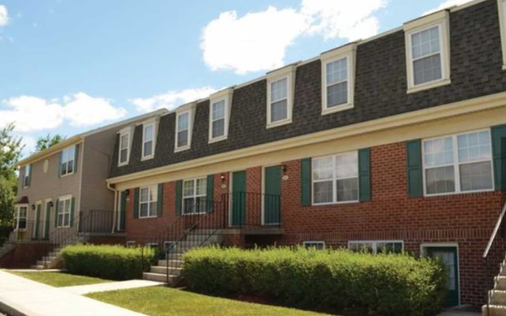 A row of two-story brick and siding townhouses with green shutters, multiple entrances, small front steps, and neatly trimmed bushes along the sidewalk on a sunny day.