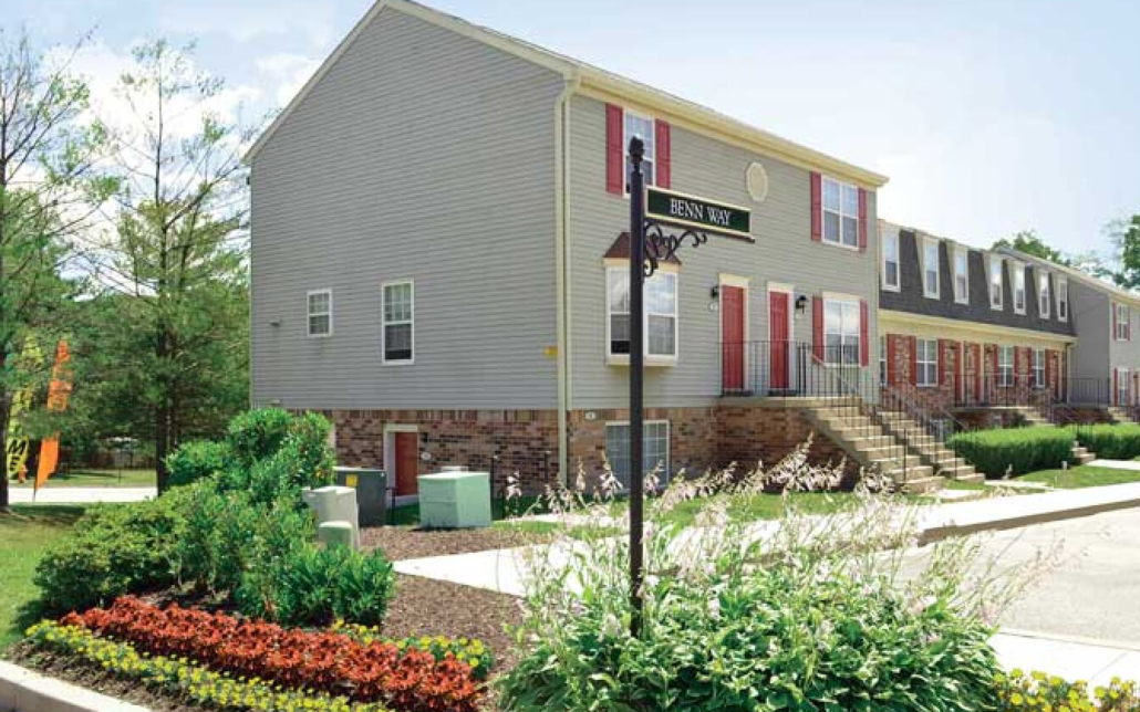 A row of gray, two-story townhouses with red doors and brick accents, situated on a clean, landscaped street with flowers, shrubs, and a street sign reading Bean Way.