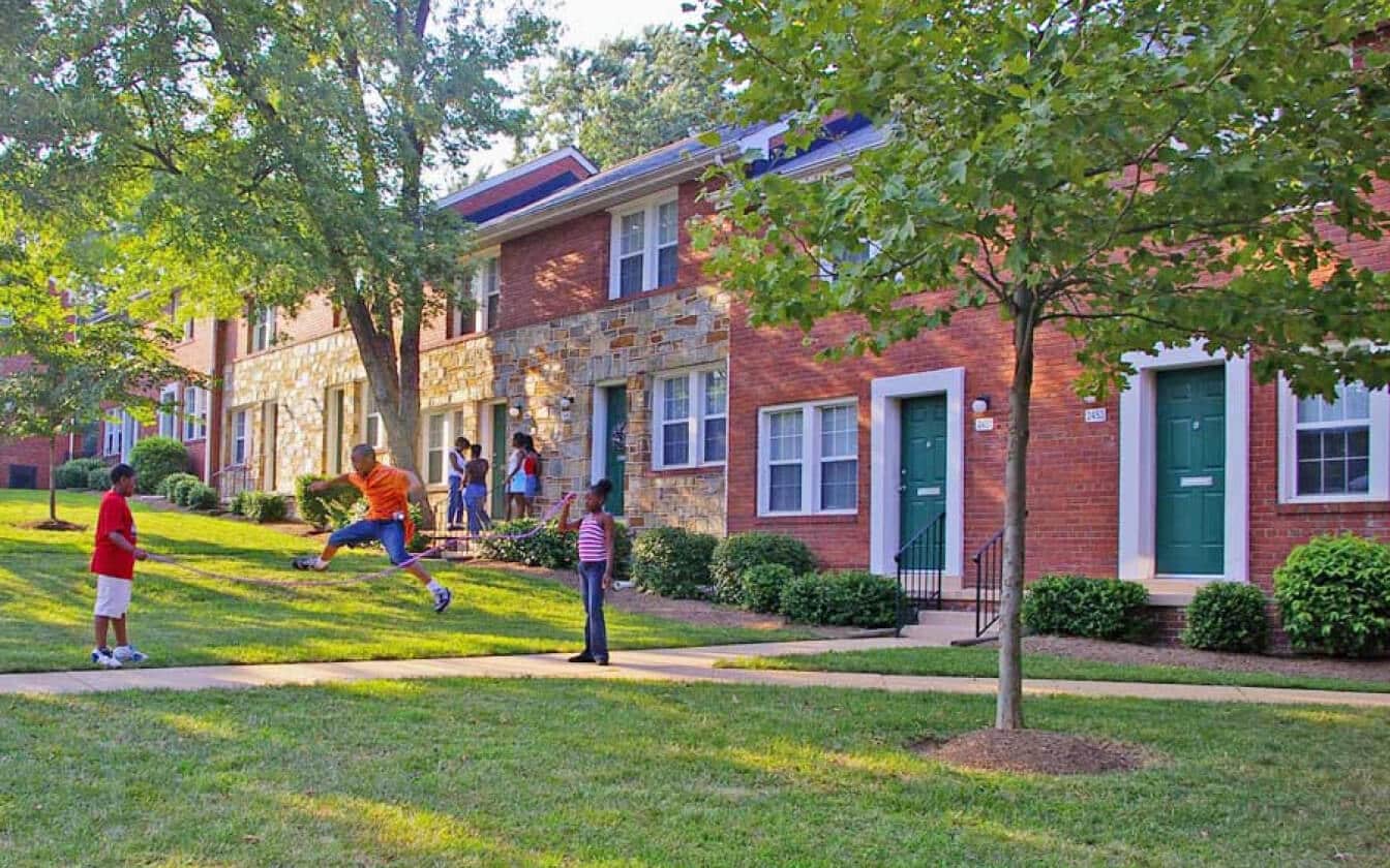 Children play on a grassy lawn in front of brick townhouses with green doors and trimmed bushes on a sunny day. One child is jumping, while others watch or stand nearby under the trees.