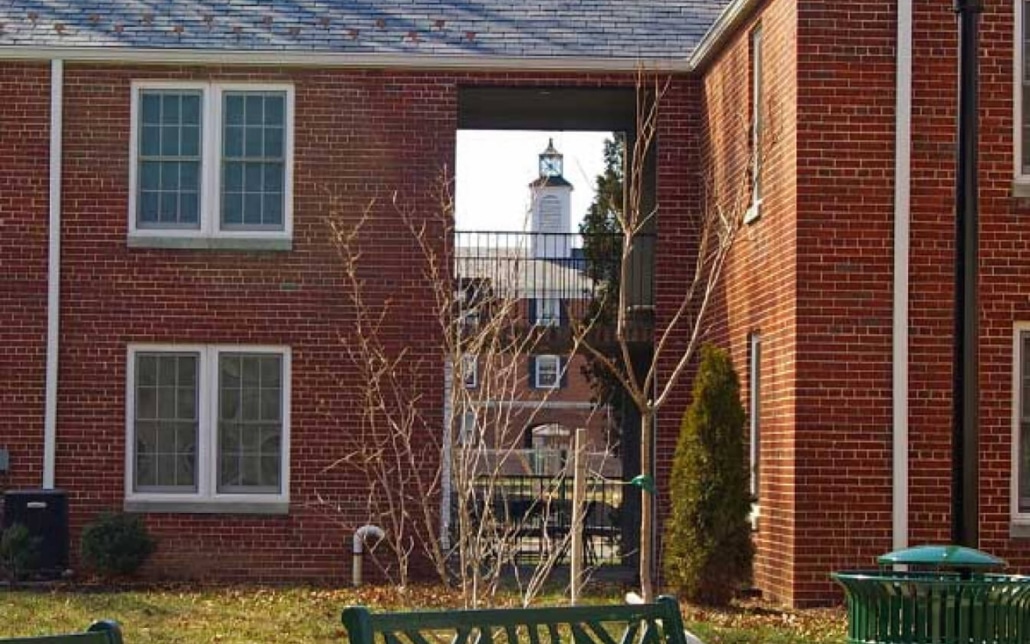 A view through two red-brick buildings reveals a distant white clock tower. Bare trees and green benches are visible in the foreground, with sunlight casting shadows on the scene.