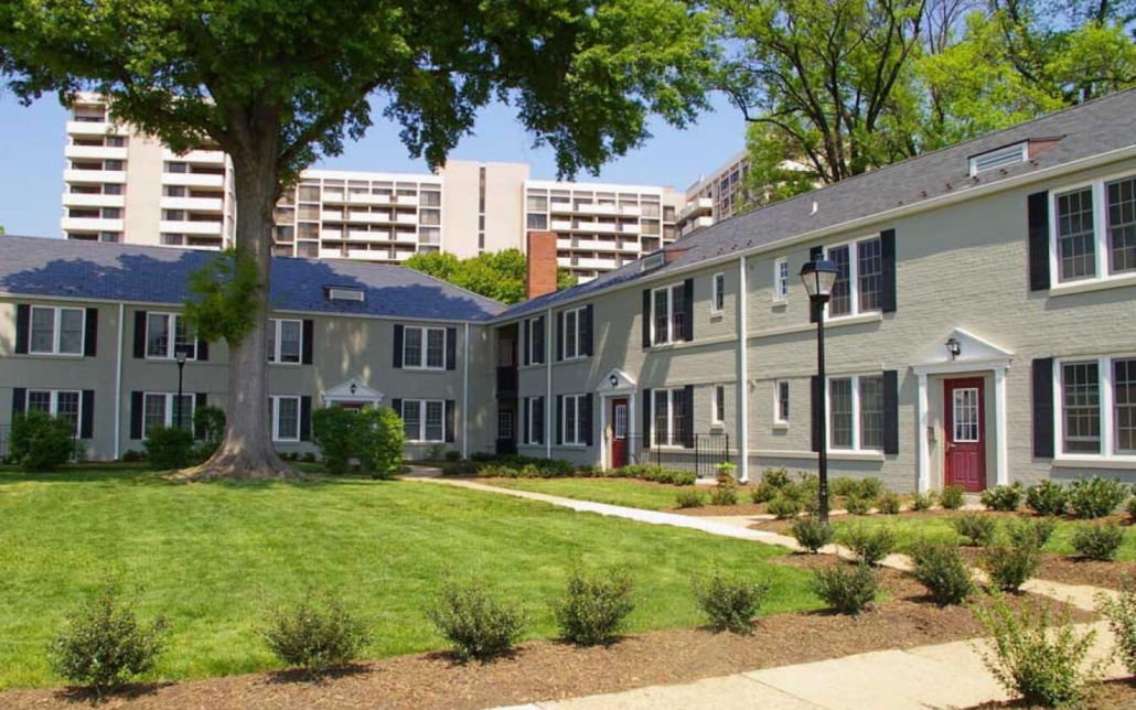 A courtyard with green grass, small bushes, and a paved walkway is surrounded by two-story gray apartment buildings with white trim and red doors. Tall trees and high-rise buildings are visible in the background.