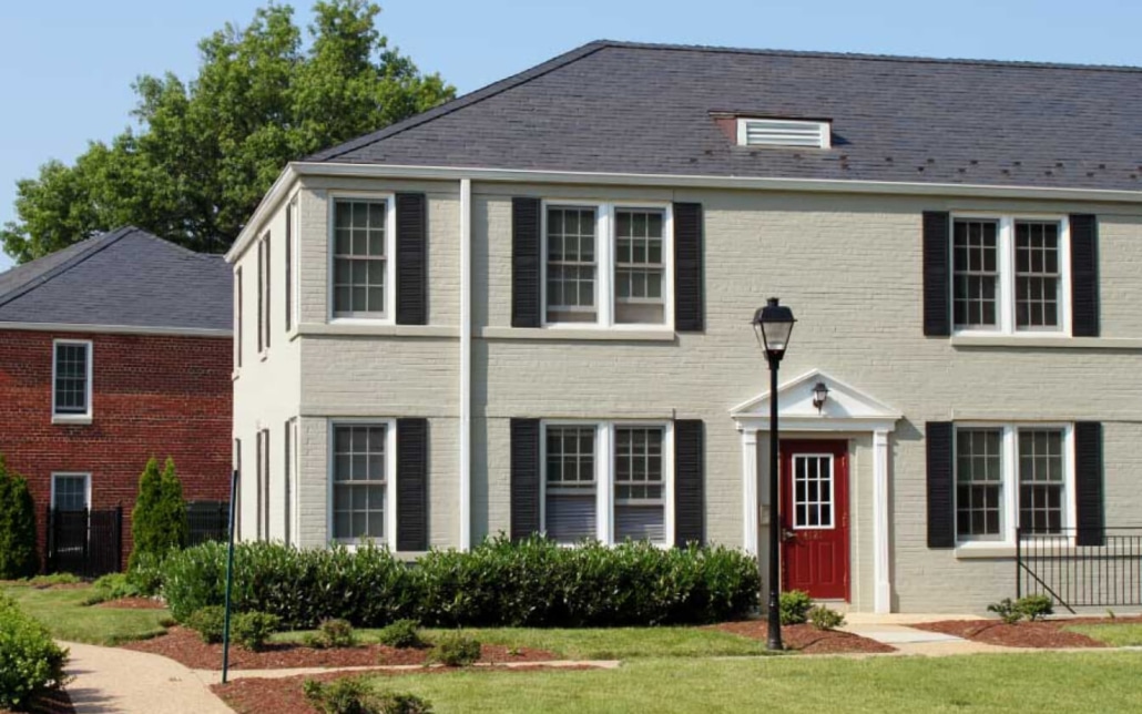 A two-story townhouse with light-colored brick, black shutters, and a red front door. There is a lamp post in front, trimmed bushes, and a sidewalk beside the building. Another brick house is visible in the background.