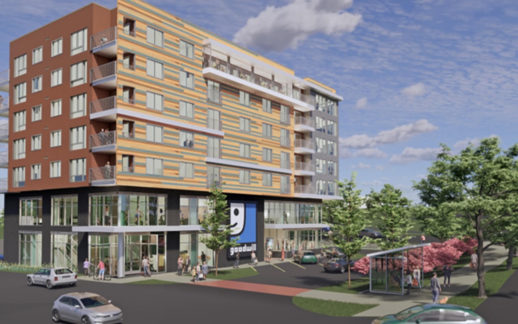 A modern multi-story building with a Goodwill store on the ground floor, people walking nearby, parked cars, trees, and a bus stop under a sunny sky with scattered clouds.