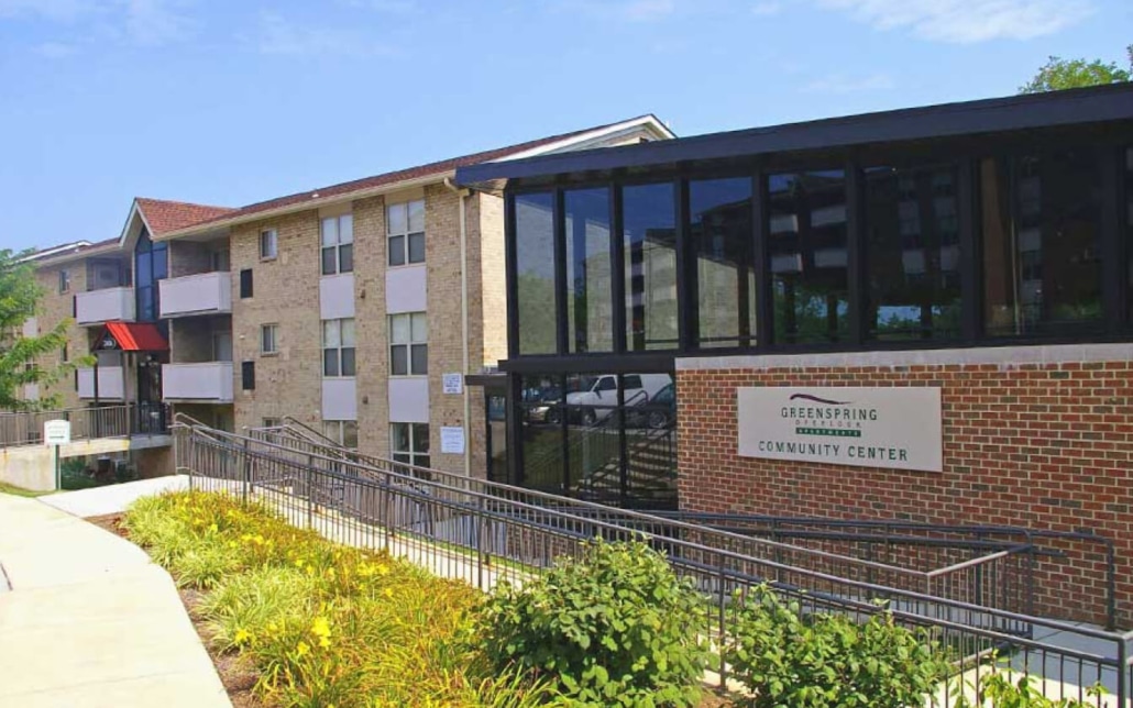 A brick and glass building with a sign reading “Greenspring Community Center,” surrounded by landscaped greenery, a walkway, and another apartment building in the background.