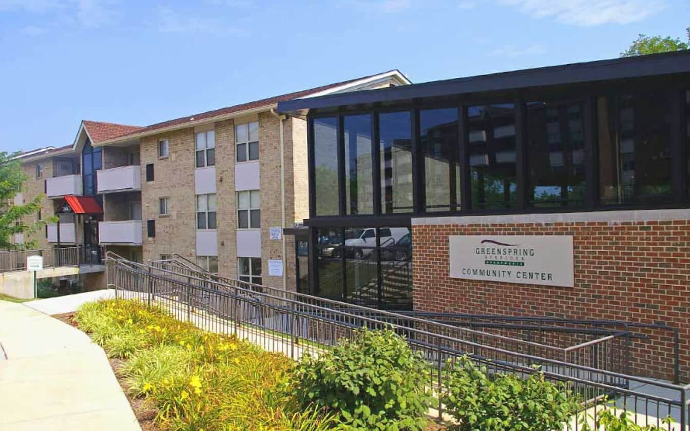 A brick and glass building with a sign reading “Greenspring Community Center,” surrounded by landscaped greenery, a walkway, and another apartment building in the background.