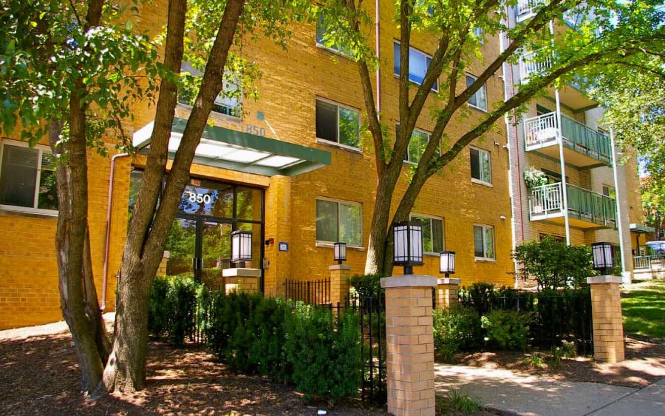 A yellow brick apartment building with balconies, green railings, and a glass entrance canopy. Trees and greenery surround the entrance, and contemporary lanterns are mounted on brick pillars along a walkway.