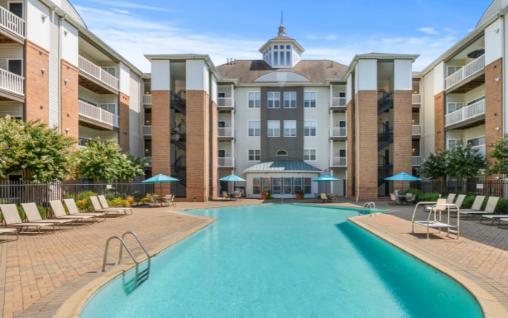 Outdoor swimming pool surrounded by lounge chairs, umbrellas, and landscaped trees in front of a multi-story apartment building with balconies and large windows under a blue sky.