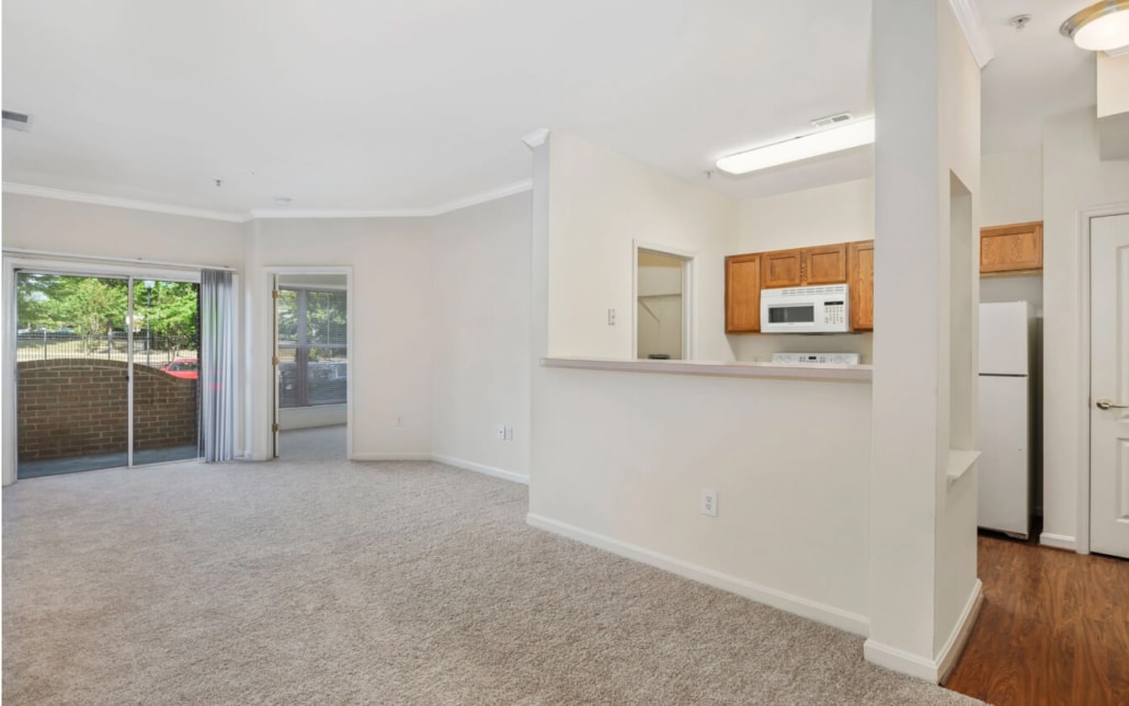 Open-concept apartment with carpeted living area, sliding glass doors to a patio, and a kitchen featuring wood cabinets, white appliances, and a pass-through counter. Bright, neutral walls and natural light throughout.