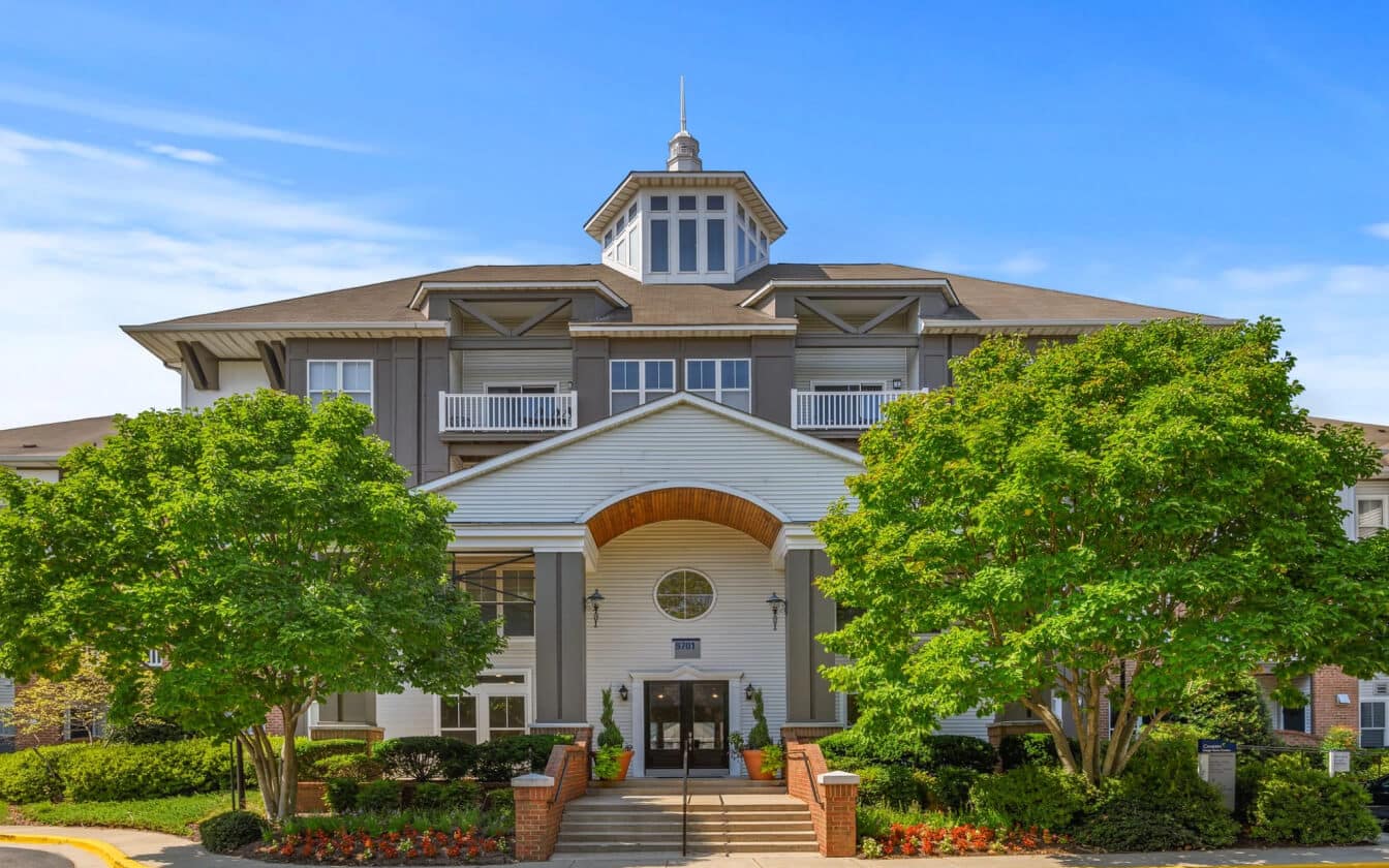 A large, modern apartment building with gray siding, white trim, and a central entrance. Green trees and neatly trimmed bushes surround the walkway leading to the front doors. The sky is clear and blue.