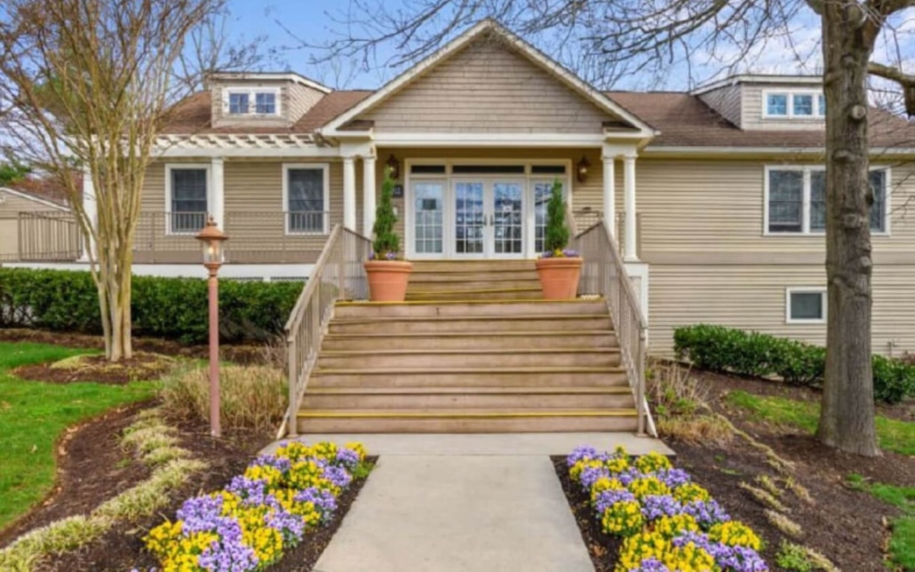 A beige house with a wooden front porch and steps, two potted plants at the entrance, flower beds with purple and yellow flowers lining the walkway, and trees on each side under a clear sky.