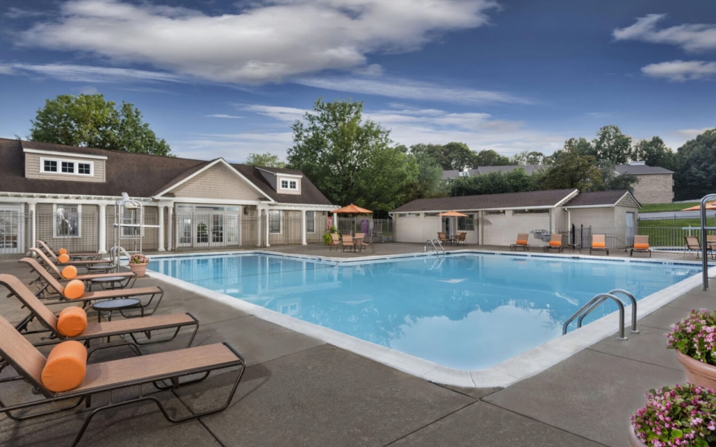 Outdoor swimming pool with clear water, surrounded by lounge chairs with orange cushions and closed umbrellas, next to a clubhouse building and greenery under a partly cloudy sky.