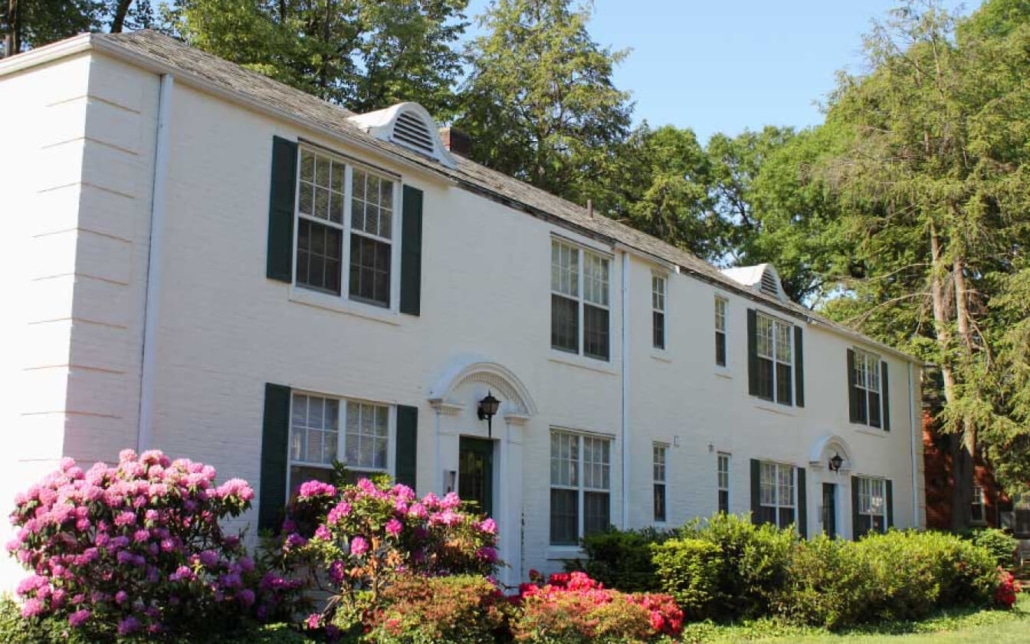 A white two-story house with black shutters, large windows, and a manicured garden featuring blooming pink and red flowers, surrounded by green shrubs and tall trees under a clear blue sky.