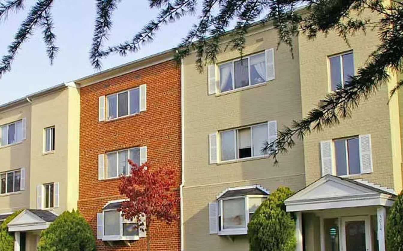 Three-story apartment building with beige and brick exterior, white window shutters, small front porches, and landscaped greenery. A tree with red leaves stands in front, and pine branches frame the top of the image.