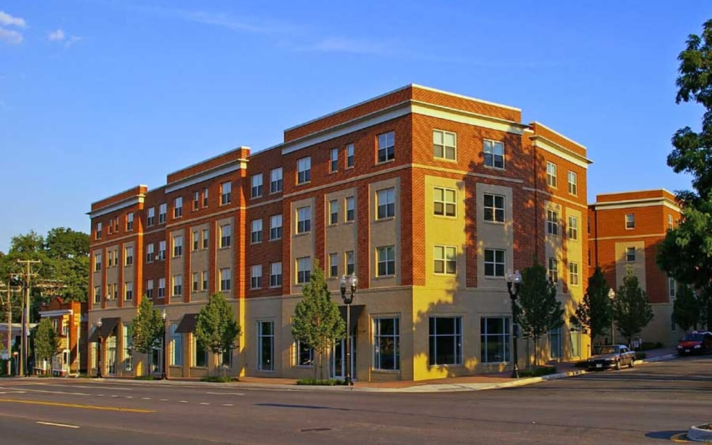 A modern four-story apartment building with red brick and beige siding, large windows, and small trees lining the sidewalk along a quiet urban street under a clear blue sky.