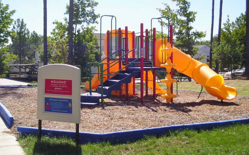 A colorful playground with slides, climbing structures, and stairs sits on mulch surrounded by a blue border. Nearby, a sign displays park information. Trees and houses are visible in the background.