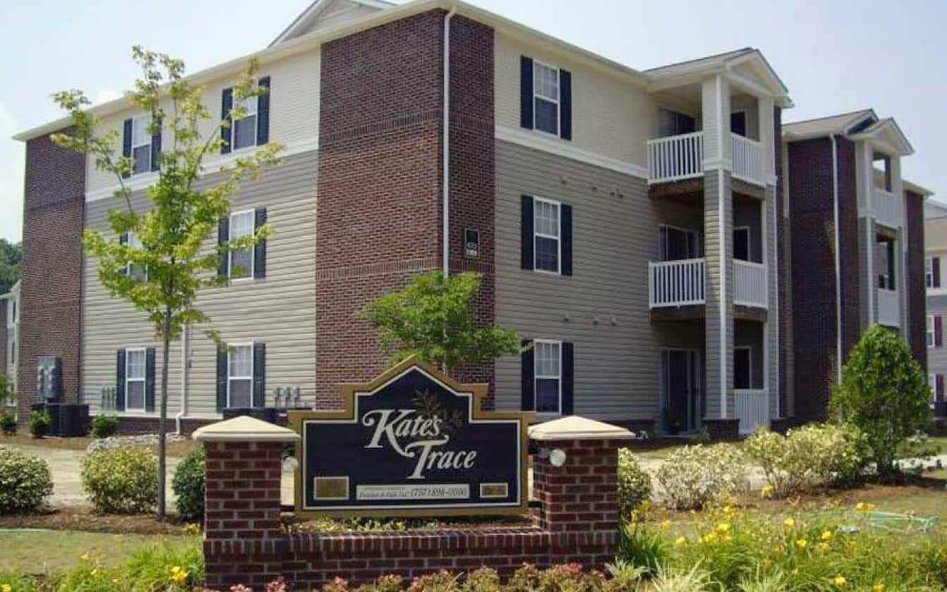 Three-story apartment building with red brick and beige siding, featuring balconies and surrounded by landscaped bushes. A sign in front reads Kates Trace.