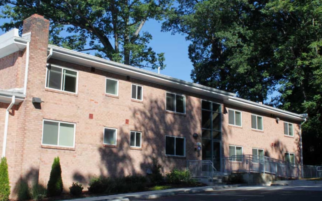 A two-story brick building with large windows and a glass entryway, surrounded by trees and greenery, on a sunny day. A ramp leads to the entrance.