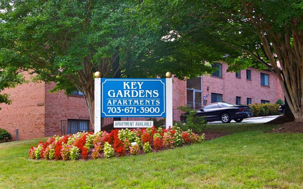 A large blue sign reading Key Gardens Apartments 703-671-3900 stands in front of a red brick apartment building, surrounded by green grass, colorful flowers, and trees. A black car is parked nearby.