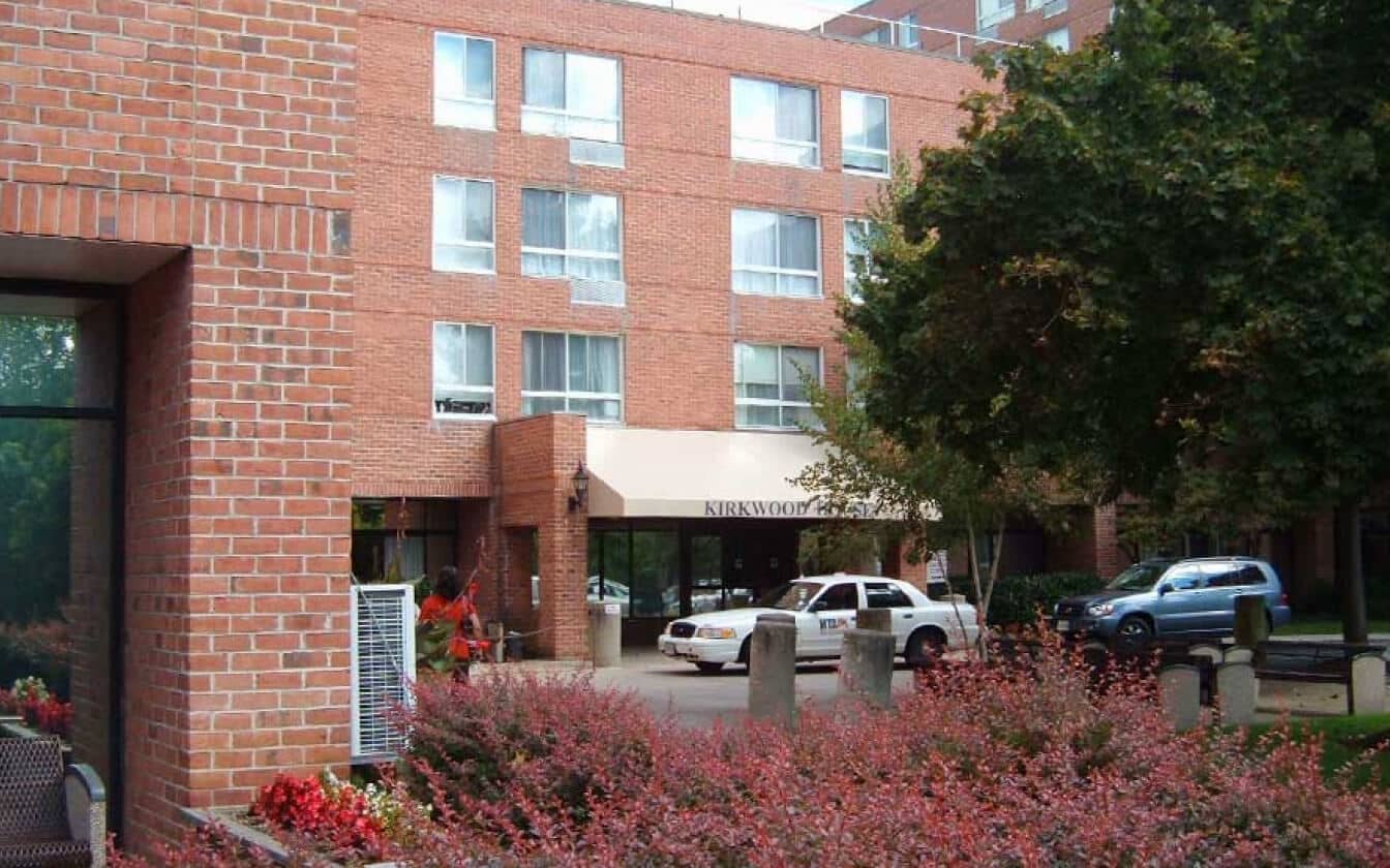 A brick apartment building with large windows, an entrance awning labeled Kirkwood, and two cars parked in front. Bushes and trees with green and red foliage are in the foreground.