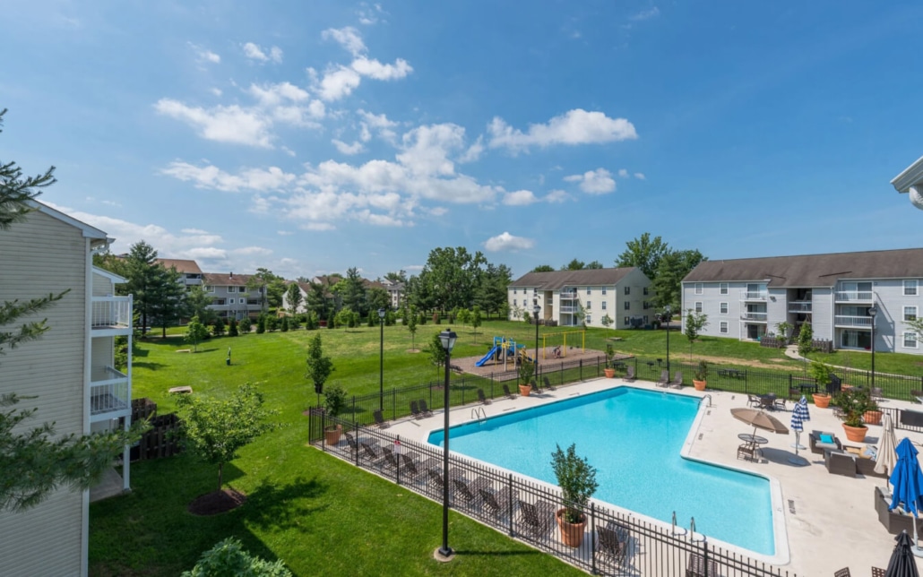 A large outdoor swimming pool with sun loungers and umbrellas is surrounded by a fence. Behind the pool are grassy fields, trees, a playground, and multiple apartment buildings under a blue sky with scattered clouds.