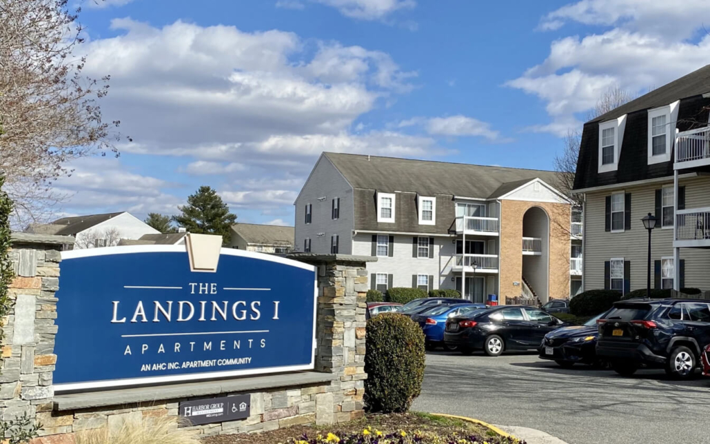 A large sign reading “The Landings I Apartments” stands in front of a parking lot with several cars and multi-story apartment buildings under a partly cloudy sky.