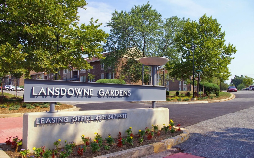 Lansdowne Gardens apartment complex entrance sign with landscaping, trees, and brick buildings in the background on a sunny day.
